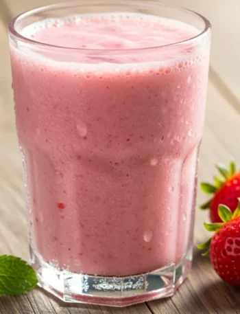 Copo de vidro com vitamina de morango cremosa e gelada, com gotículas de condensação escorrendo e morangos frescos ao lado sobre uma mesa de madeira.